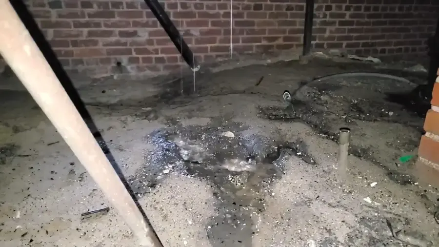 Wet basement floor with standing water along a brick foundation wall and a drain pipe